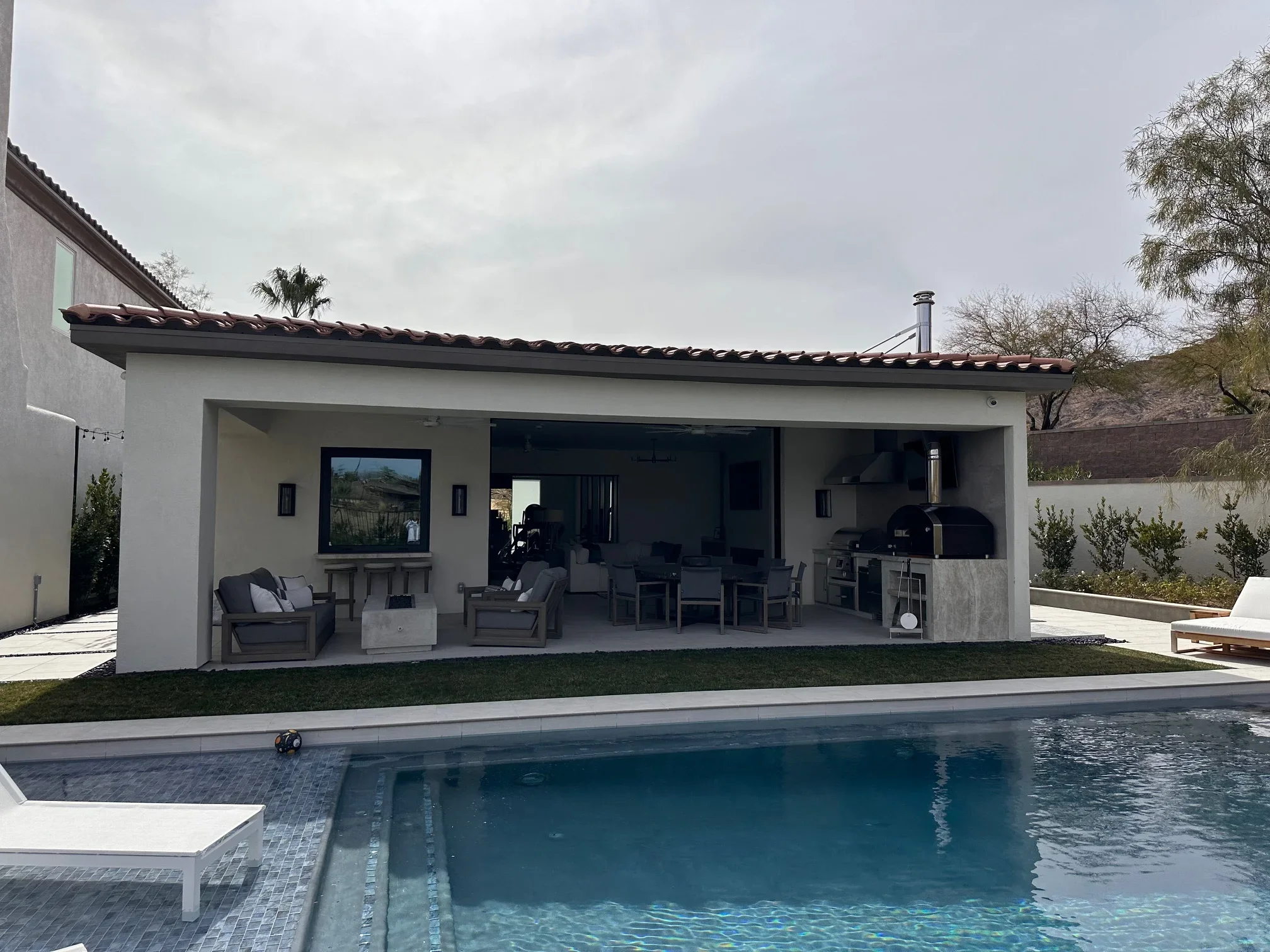 Modern white stucco pavilion with metal roof, outdoor kitchen, pizza oven, and pool deck