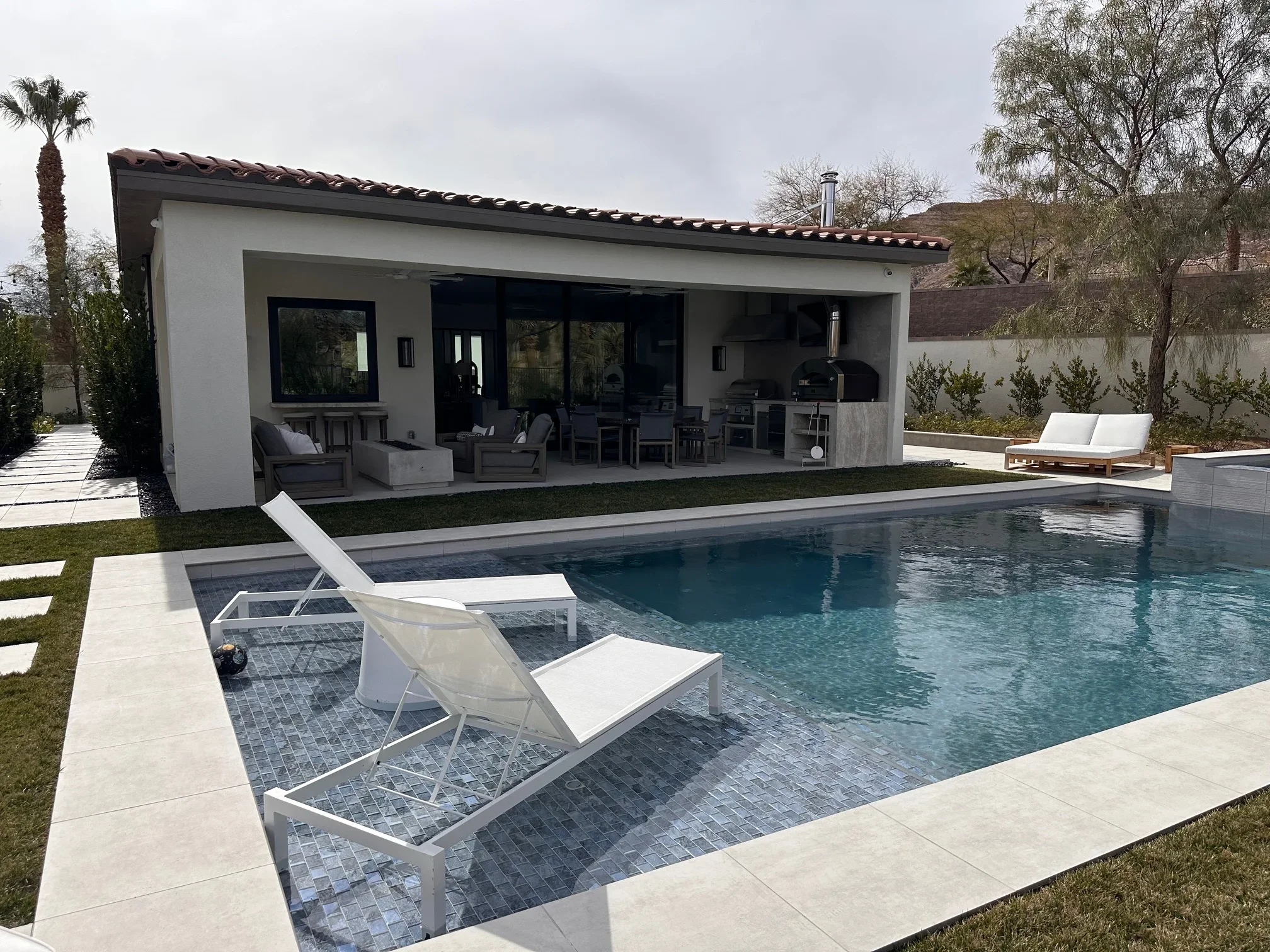 Modern white stucco pool house with black glass doors, Spanish tile roof, and resort pool with mosaic tile accents