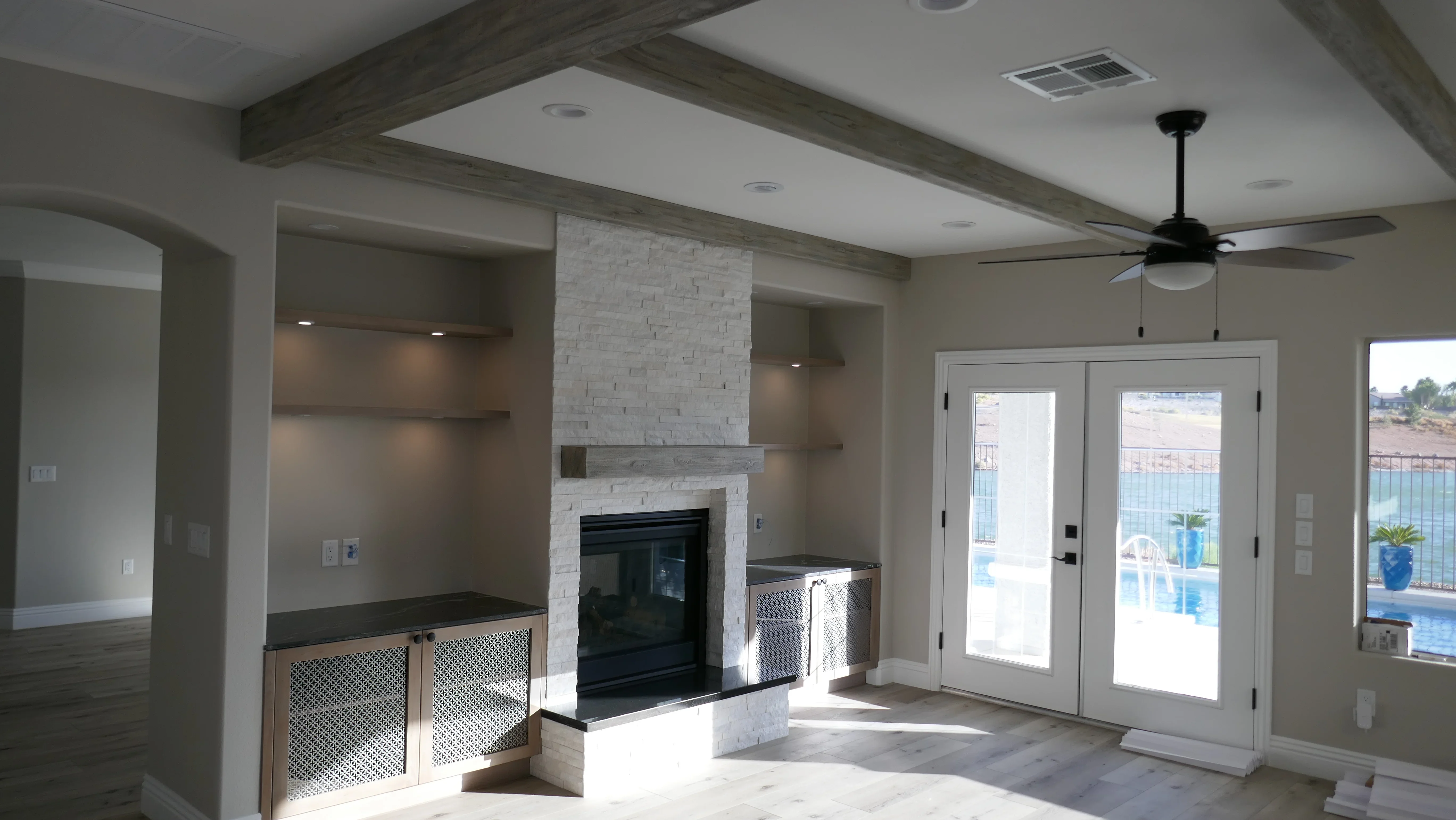 Great room with white brick fireplace, built-in shelving, wood beams, and French doors to pool