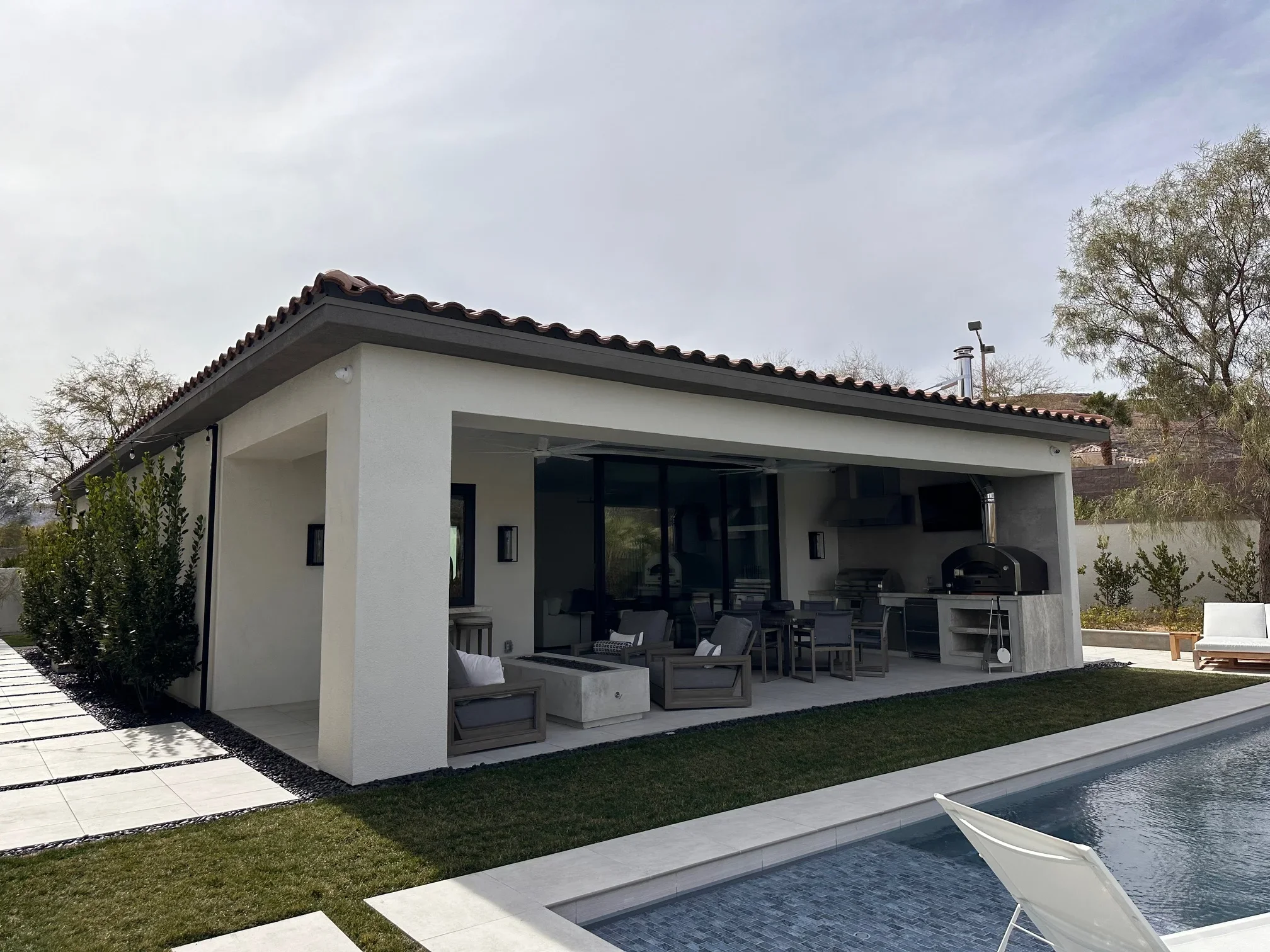 White stucco pool house with tile roof, sliding glass doors, and covered patio overlooking pool