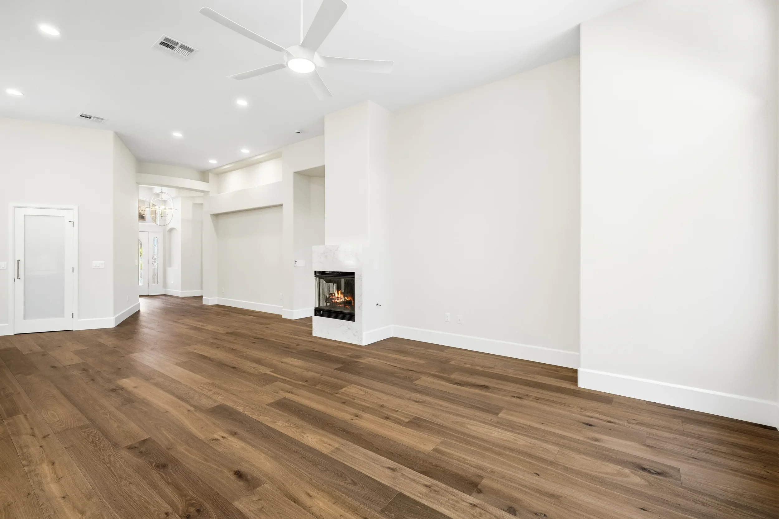 Open living room with wood flooring, white walls, recessed lights, ceiling fan, and built-in fireplace