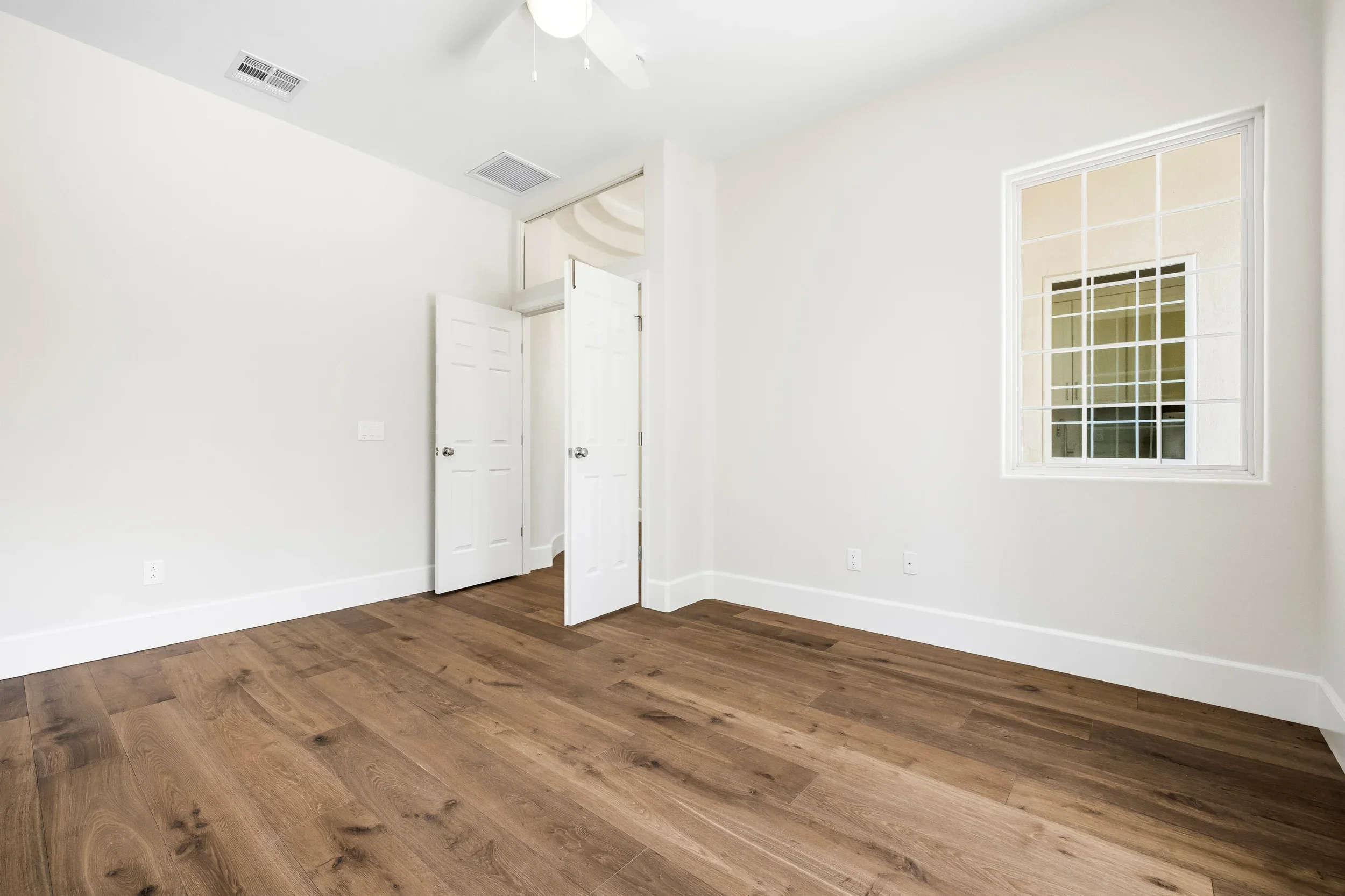 Empty bedroom with hardwood flooring, white doors, and large window with white trim