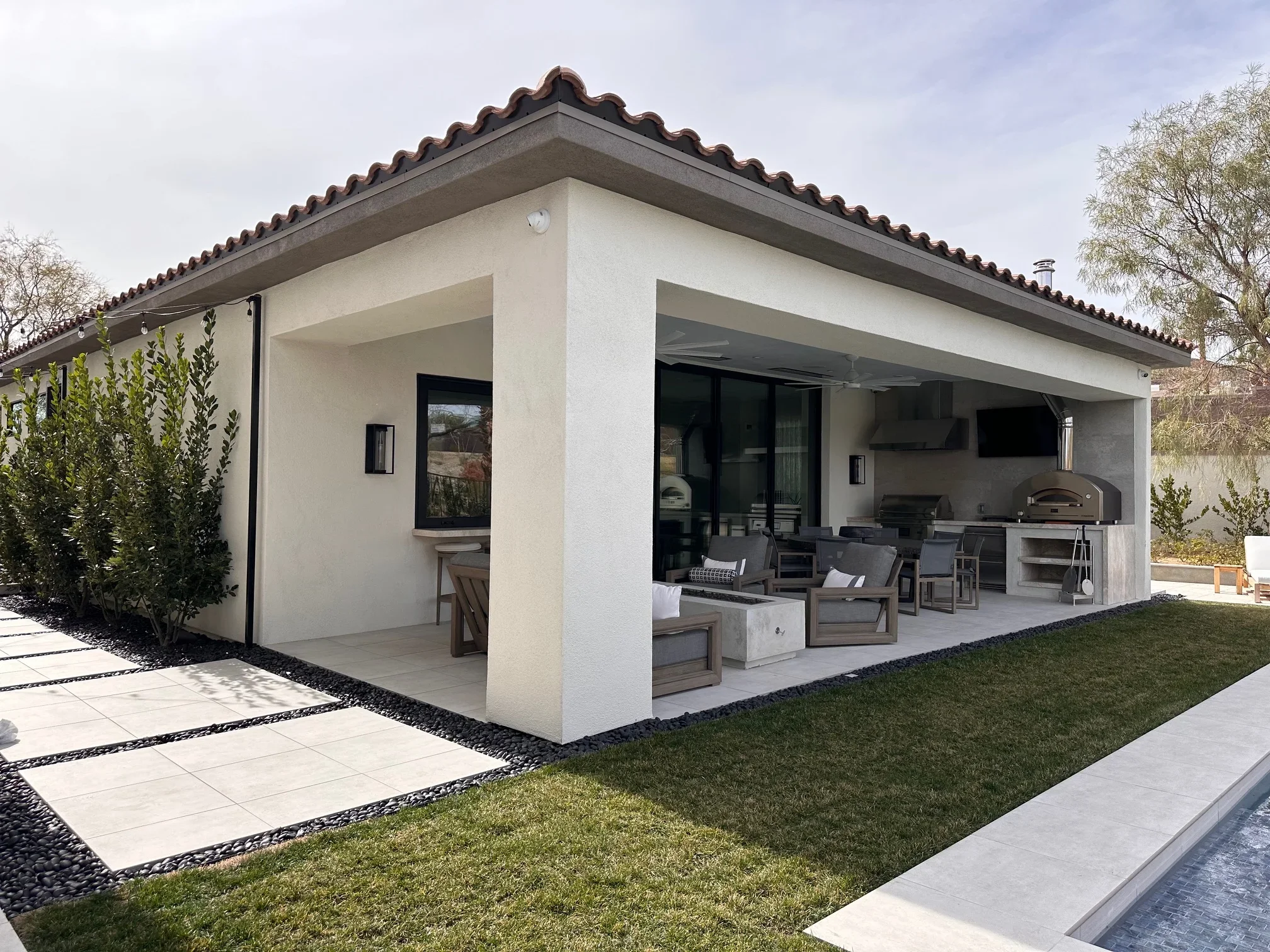 Modern stucco pool house with clay tile roof, outdoor kitchen, and covered patio seating area