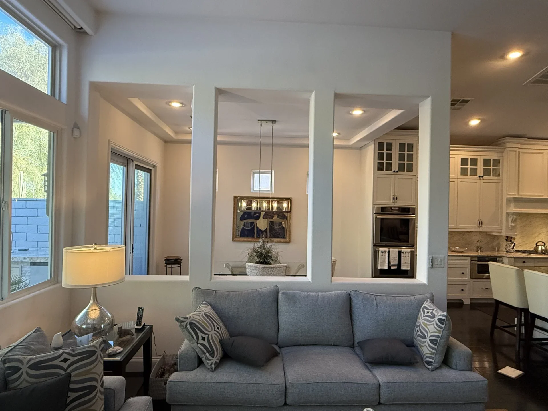 Open concept living room with white columns, recessed lighting, gray sofa, and kitchen with cream cabinetry visible beyond